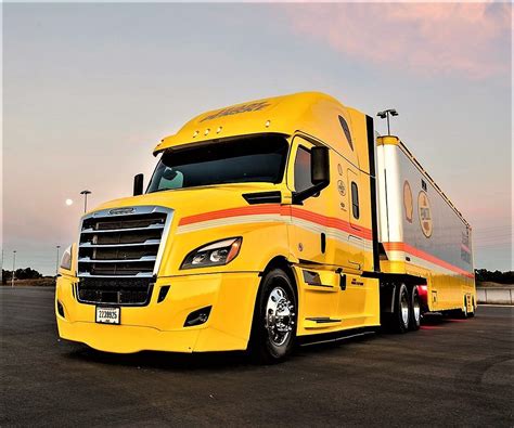 A Large Yellow Semi Truck Parked In A Parking Lot At Dusk With The Sun
