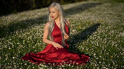 Blondynka w czerwonej sukience siedzi na łące pośród stokrotek