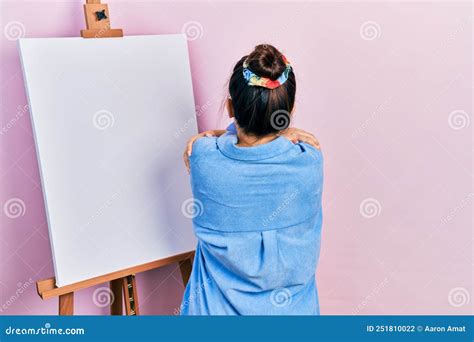 Young Hispanic Woman Standing By Painter Easel Stand Hugging Oneself