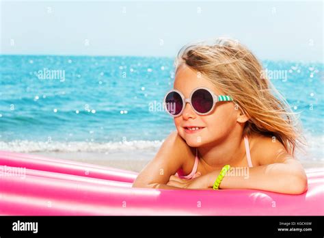 Portrait Of Little Blond Girl In Sunglasses Sunbathing On Pink Inflatable Lounge Against Blue