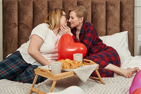 A Lesbian Couple In Pajamas Lies On The Bed In Front Of Them Breakfast On A Folding Table And A