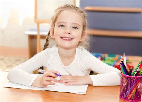 Cute Little Girl Is Writing At The Desk In Preschool Stock Photo