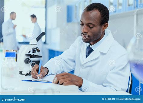 African American Scientist In White Coat Taking Notes While Working In