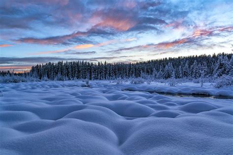 Snowy Mounds And Conifer Forest With Sunset Illuminating The Sky Over