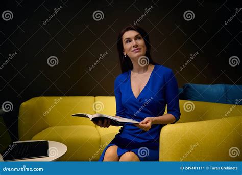 Delightful Brunette Woman In Blue Dress Sits On Yellow Sofa Holds Catalog Looks Away Dreamily