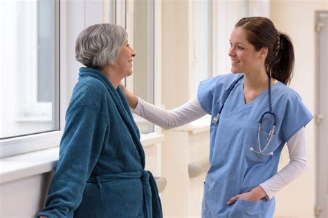 Smiling Female Nurse Caring For Senior Patient In Hospital Corridor