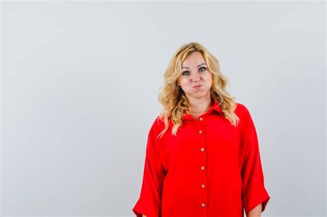 free photo blonde woman standing straight puffing cheeks in red blouse and looking happy