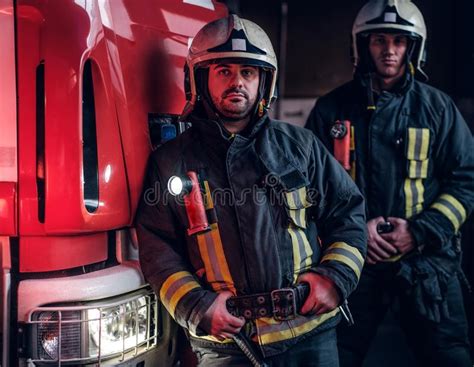 Two Firemen Wearing Protective Uniform Standing Next To A Fire Truck In