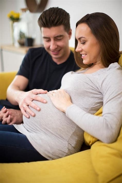 Pregnant Couple Sitting On Sofa With Man Touching Womans Stomach Stock