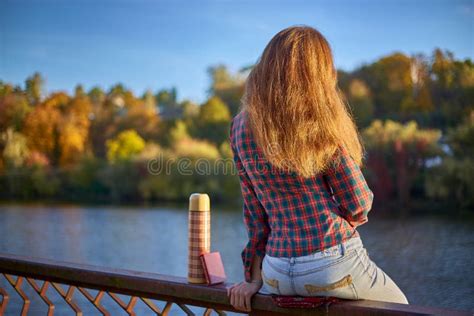 Young Girl In Plaid Shirt Sitting On Iron Railing At River Embankment