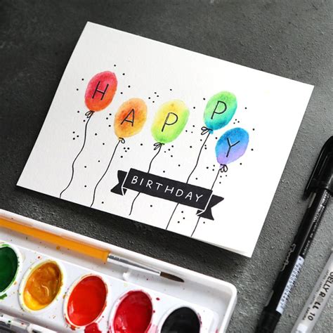 A Happy Birthday Card With Balloons And Watercolors On The Table Next To It