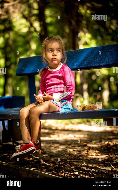 Beautiful Little Girl Sitting On A Park Bench On A Summer Day Eating