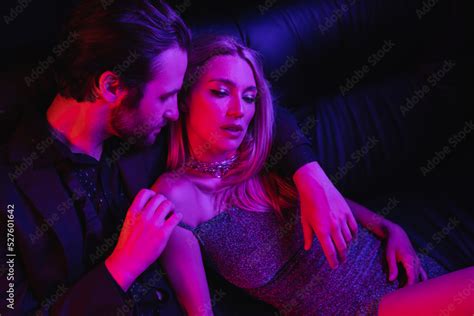 sexy man undressing blonde girlfriend in dress on leather couch with lighting isolated on black