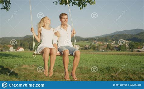 Portrait Young Sunlit Couple Sitting On Wooden Swing And Gazing Into