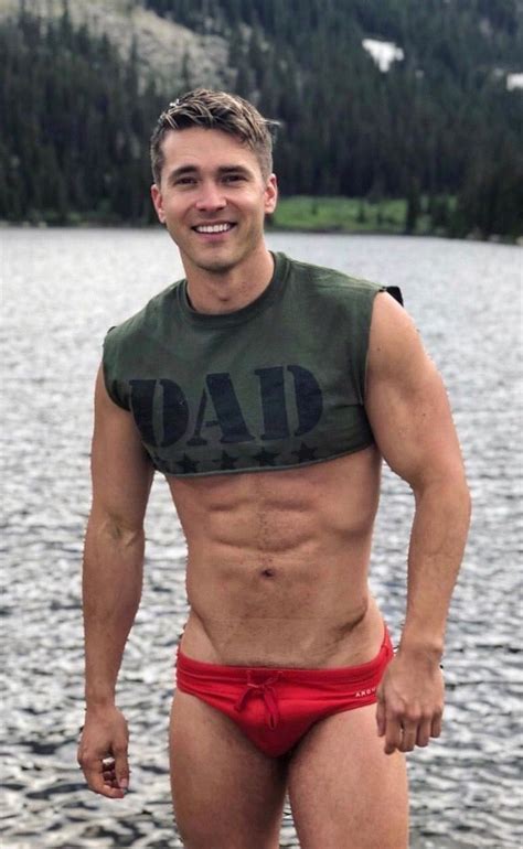 A Man With No Shirt On Standing In Front Of A Body Of Water Wearing Red Trunks