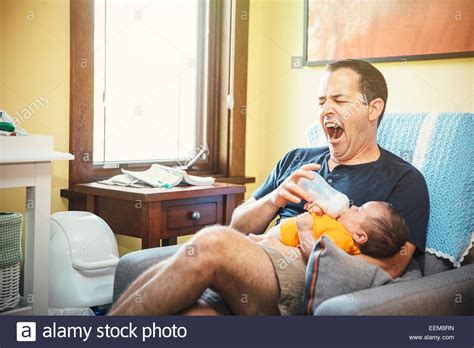 Yawning Caucasian Father Feeding Baby Boy In Living Room Stock Photo