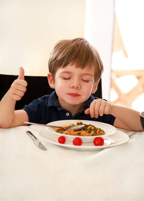 Hungry Child Sitting In Chair At Table In Kitchen And Eating With Spoon