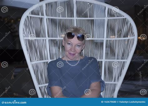 a mature woman in sunglasses on her forehead is smiling while sitting on a wicker chair stock