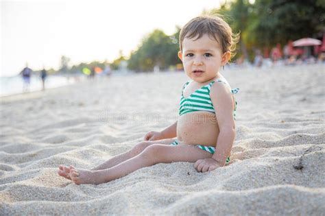 Cute Toddler In A Swimsuit Looks At Camera And Sits On A Sandy Tropical