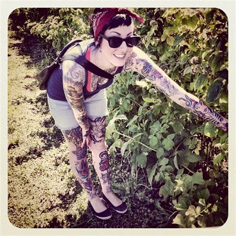 a woman with lots of tattoos on her arm and leg standing in front of bushes