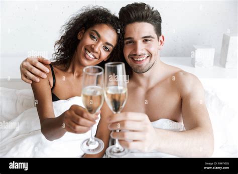 Beautiful Happy Young Multiethnic Couple Relaxing In Bed Under Blanket Drinking Sparkling