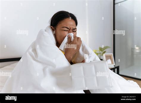 Sick Woman Sitting On Bed Wrapped In A Blanket Blowing Nose In A Tissue
