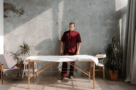 Male Masseur Wearing Red Uniform Standing Near Table By Stocksy