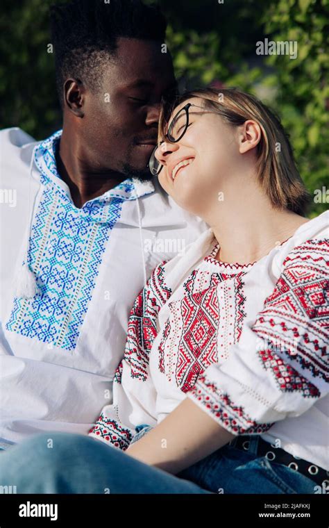 interracial happy couple sits on bench in garden dressed in ukrainian traditional ethnic