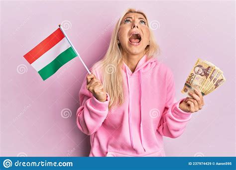 Young Blonde Woman Holding Hungary Flag And Forint Banknotes Angry And Mad Screaming Frustrated