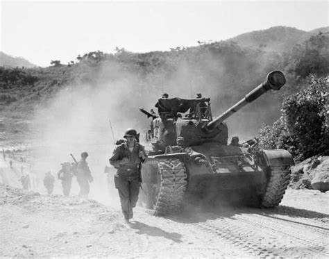 An Army Tank Driving Down A Dirt Road Next To Other People On The Side