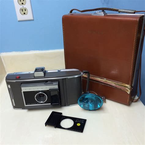 An Old Fashioned Camera Next To A Brown Leather Case On A White Table With A Blue Glass
