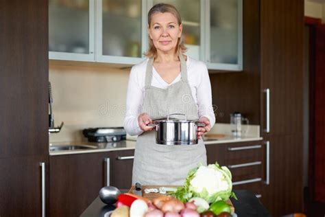 Mature Woman In The Kitchen Prepares Food And Holds A Pot Of Soup