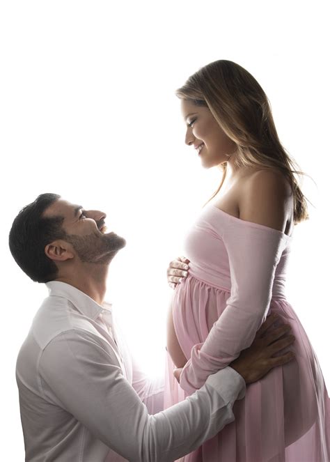 A Pregnant Couple Standing Next To Each Other In Front Of A White Background And Looking At Each